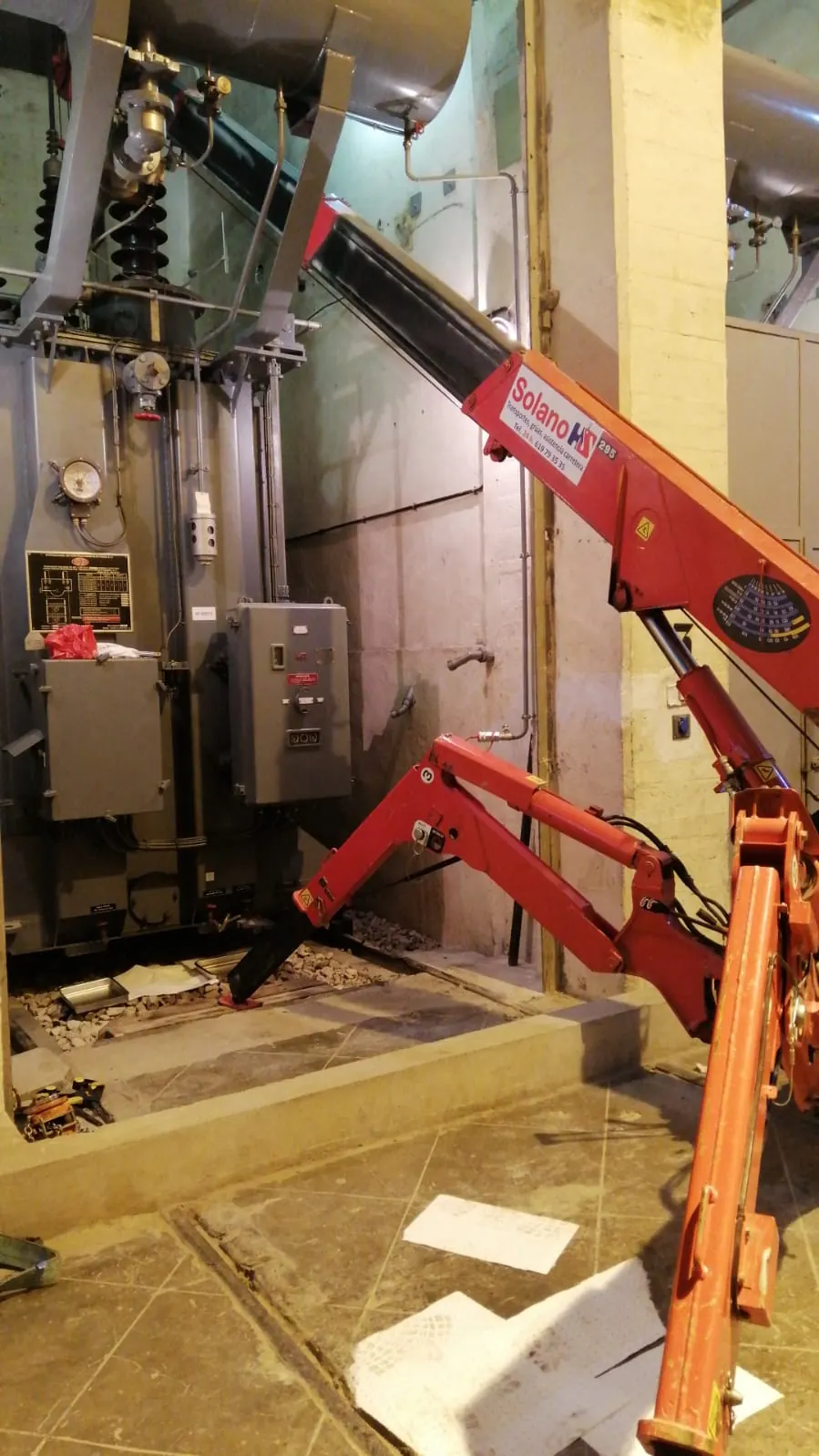 Mini grúa sobre orugas trabajando dentro de una sala técnica, entre un transformador y la pared